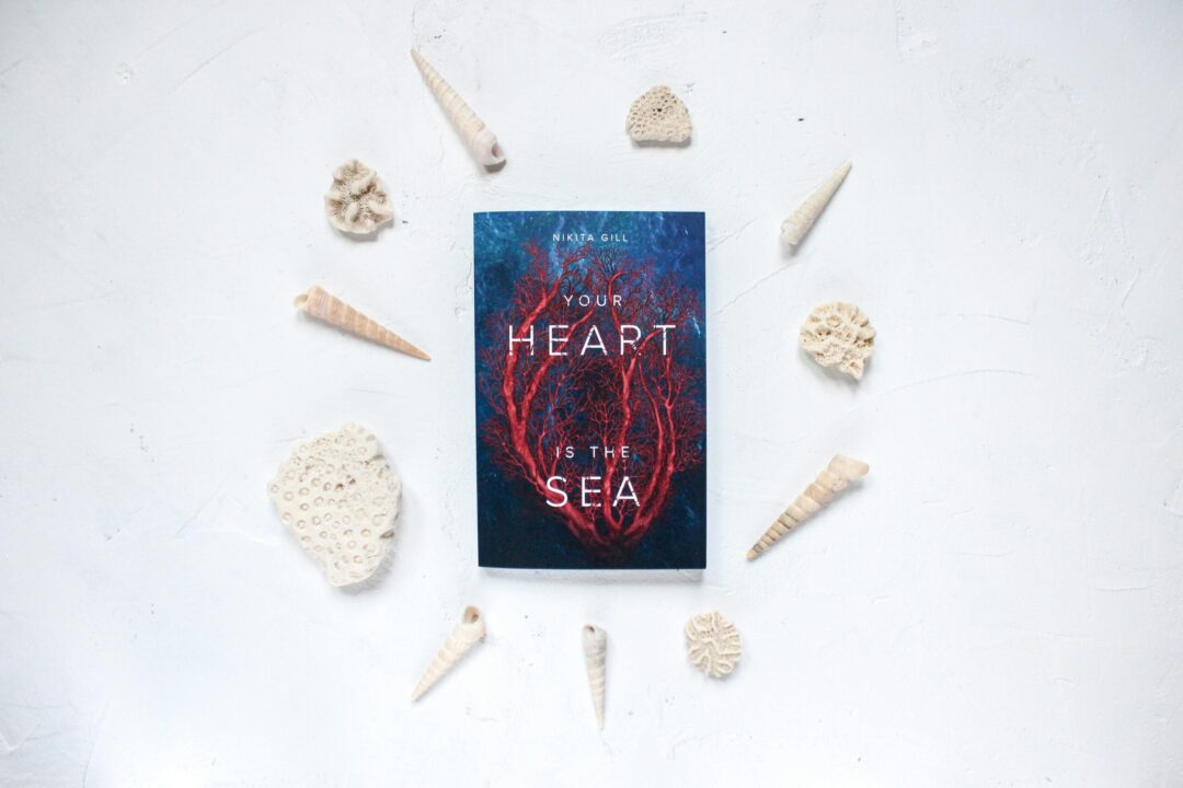 Flatlay featuring a book titled 'Your Heart is the Sea' surrounded by seashells on a white background.