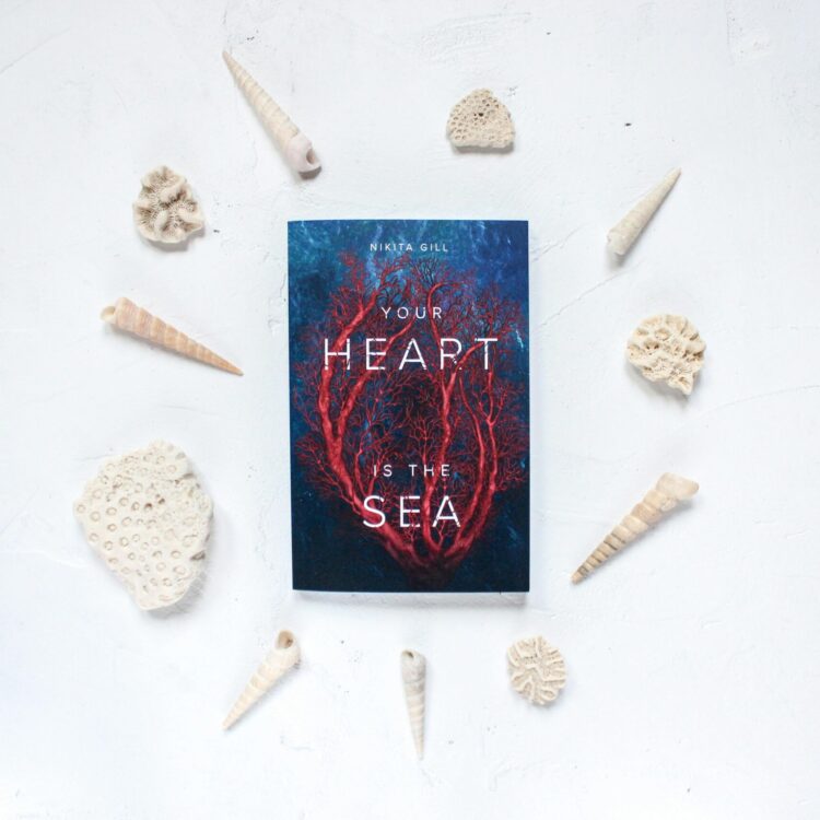 Flatlay featuring a book titled 'Your Heart is the Sea' surrounded by seashells on a white background.