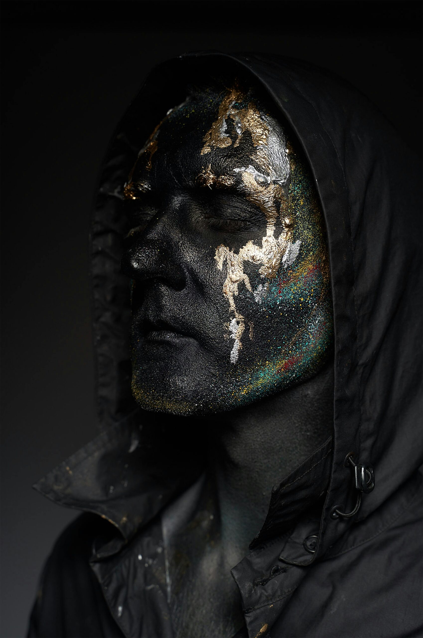 Close-up of a man with glittery face paint wearing a dark hoodie, expressing an eerie artistic style.